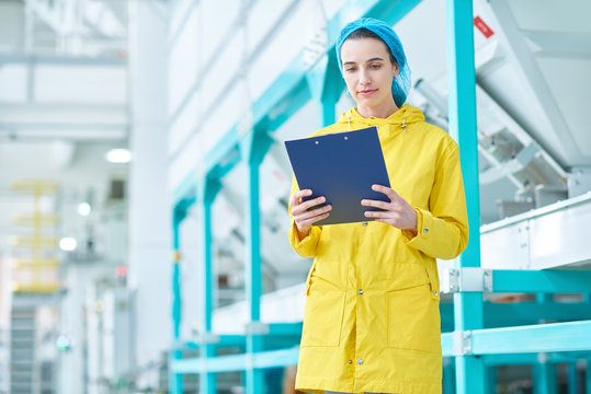 Portrait of cheerful young woman holding clipboard standing in clean production workshop of modern plant, copy space