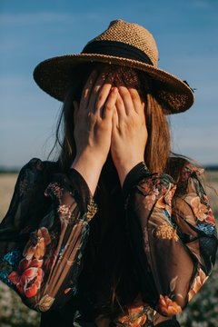 Sunny portrait of young woman hiding her face with hands and wearing straw hat