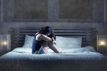 depressed Latin girl sitting on bed thoughtful and worried about problem or feeling sick and unwell...