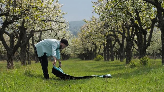 A young man puts a blanket on the grass in the park for a romantic dinner with a girlfriend