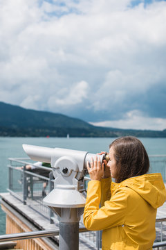 Girl in a yellow raincoat looking through coin-operated telescope at the opposite bank of the lake in Alps