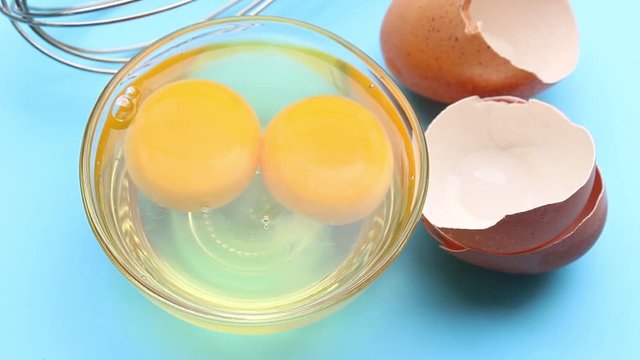 Raw egg in glass bowl with eggshell and whisk on blue background