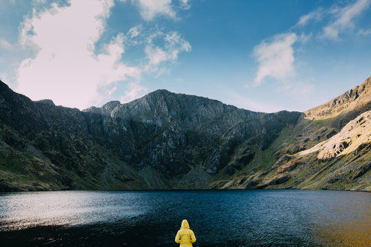 A person in a yellow jacket in the middle of the frame, looking towards a mountain peak and a lake