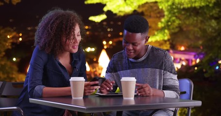 Attractive black male and female using tablet computer outdoors at night, Friends meeting for coffee using modern technology and talking, 4k - Powered by Adobe