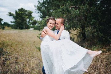 Beautiful and smiling newlyweds gently embrace in a green pine forest on a sunny day. The young groom hugs and kisses from behind a beautiful bride in a lace dress in nature. 