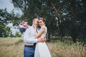 Beautiful and lovely newlyweds are hugging in a green pine forest on a sunny day. A wedding portrait of a young groom and a beautiful bride in a lace dress in nature. Smiling newlyweds.