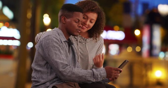 Cute African American couple have fun on the Champs-Elysees, Happy female sits on her boyfriend's lap while he texts on smartphone, 4k