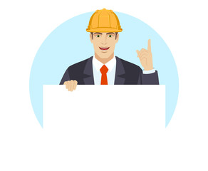 Businessman in construction helmet holding white blank poster and pointing up
