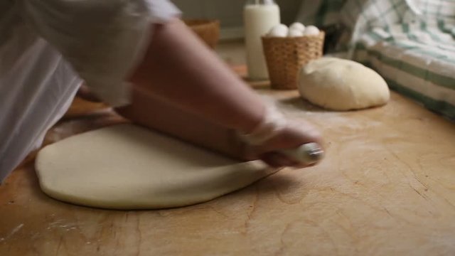 Woman baker rolls the dough with a rolling pin on a background of ingredients on a wooden table and flour, close-up