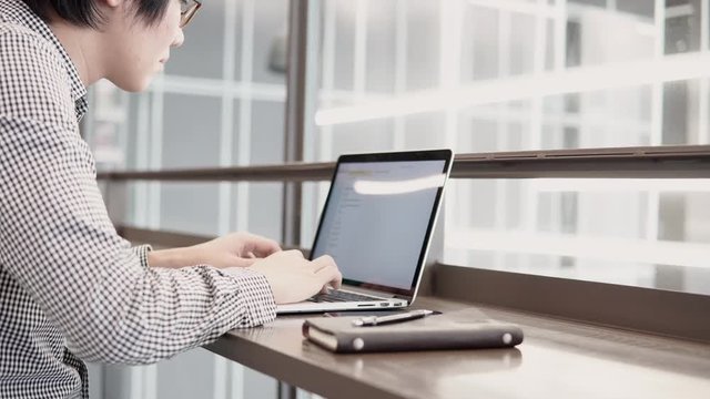 Young Asian business man using laptop computer in working space with smartphone and notebook on wooden desk. Male hand typing on laptop keyboard. Freelance lifestyle in digital age concept. 4K video