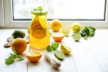 Iced lemonade pitcher, wooden juicer, cold citrus infused water with lemon & lime slices, mint leaves, cutting board on white wooden windowsill. Apartment window background, close up, copy space. © Evrymmnt