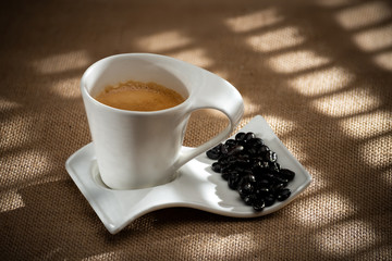 white cup of coffee with coffee beans on a textured sack background with lights and shadows