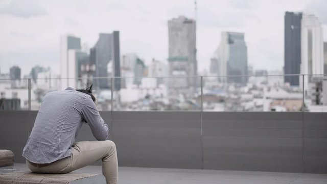Young Asian business man feeling stressed and frustrated with job and life problems sitting on bench on office building rooftop terrace. city view in background. Major depressive disorder concept.