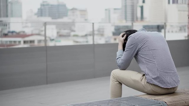 Young Asian business man feeling stressed and frustrated with job and life problems sitting on bench on office building rooftop terrace. city view in background. Major depressive disorder concept.