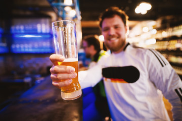 Joyful football fan is holding his beer pint up and saluting while sitting at the bar.