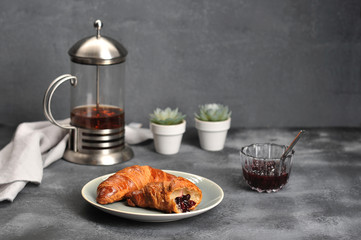 Two crispy croissants on a plate. One croissant is broken and smeared with jam. Next a cup of jam and a teapot with tea. In the frame, cloth napkin. Gray background. Close-up.