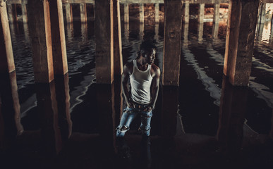 Young African man stands in water under bridge on background of concrete supports.