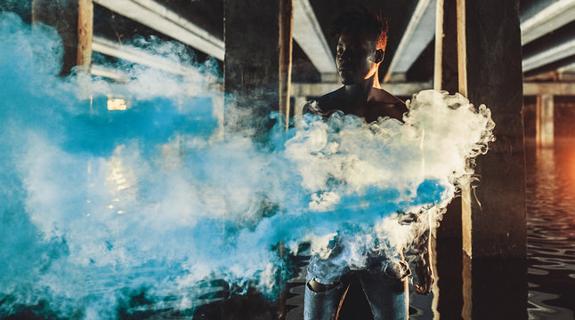 Young African man stands under bridge and holds colored blue smoke bomb in his hands. - Powered by Adobe