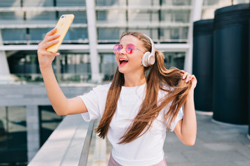 Modern pretty happy woman with nude make up, long hair, wears white t-shirt and pink glasses making selfie outdoor in morning sinshine with true happy smile