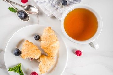 Sweet summer dessert, homemade baked mini croissants with berry jam, served with tea, fresh raspberries, blueberries and mint. On a white marble table, copy space top view