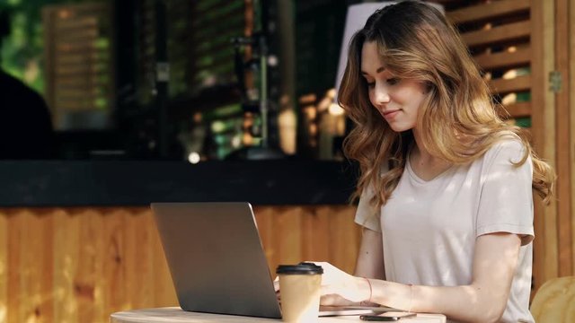 Pleased brunette woman in casual clothes sitting by the table while using laptop computer and drinking coffee at cafe outdoors
