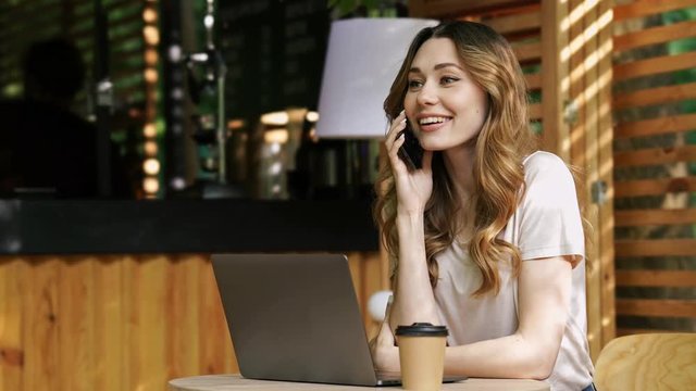 Smiling brunette woman in casual clothes sitting by the table with laptop computer while talking by smartphone at cafe outdoors