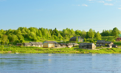 Fototapeta na wymiar The northern village is on the banks of a river on background of blue sky. It is a landscape with wooden houses near a forest. There are clear, warm, sunny.
