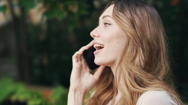 Side view of Pleased brunette woman in casual clothes talking by smartphone while sitting on bench outdoors
