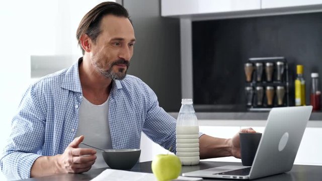 Side view of Pleased mature man in shirt sitting by the table with laptop computer while having breakfast at home