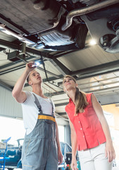 Low-angle view of a reliable auto mechanic holding a flashlight, while checking the parts of the lifted car of a woman in a modern automobile repair shop