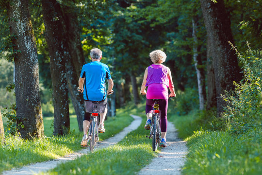 Full length of a happy and active senior couple wearing cool fitness outfits while riding bicycles outdoors in the park