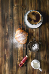 Homemade croissant with sugar powder, cup of coffee, jug of milk, vintage sieve over wooden plank background. Flat lay, space.