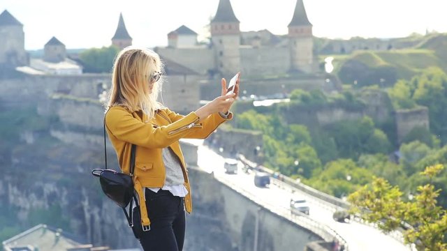 fashionable blonde girl taking photo selfie in old town standing against of historic brick castle on the top of a cliff female tourist using smartphone in old european town traveling cultural heritage