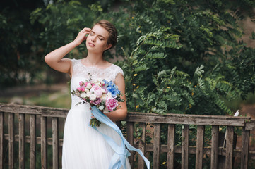 A beautiful and sweet bride in a lace dress stands against the background of an old hut in the village and a wooden fence with green foliage and straightens her hair.