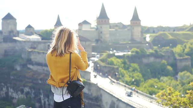 young fashionable female tourist using smartphone mobile for taking a photo of beautiful ancient old brick castle in summer sunny windy day back view technology device entertainment traveling concept