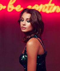 Fashion portrait of young beautiful woman with middle length dark hair wearing green sequins dress posing against red wall with neon letters in night bar