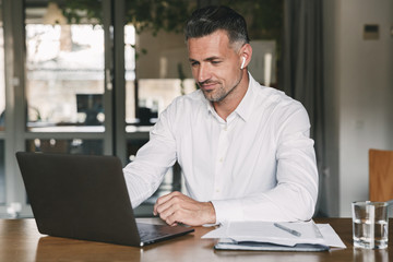 Image of handsome business man 30s wearing white shirt sitting at table in office, and working at laptop using wireless earpod