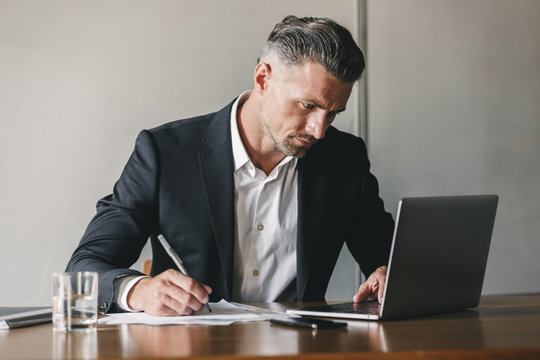 Image Of Concentrated Confident Businessman 30s Wearing White Shirt And Black Suit Working At Laptop In Office, While Writing Down Notes