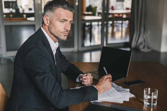 Image of concentrated confident businessman 30s wearing white shirt and black suit sitting at table in office, during work with documents and laptop