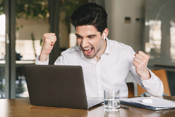 Obraz premium Portrait of successful businessman 30s wearing white shirt and bluetooth earbud screaming and clenching fists, while looking at laptop in office