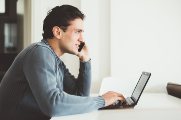 Handsome young man sitting indoor talking smart phone using computer - business, technology, internet concept