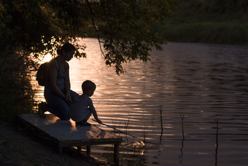 a young mother with a child is standing by the river, looking at the beautiful Sunset.