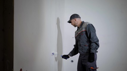 professional painter is wearing working uniform and cap is painting wall by roller with white color, side view