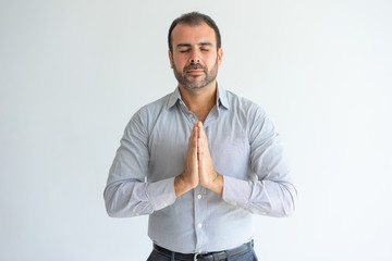 Serene middle-aged Latin businessman in shirt meditating alone. Calm concentrated handsome man with beard relaxing and holding hands in Namaste. Unity of soul and body concept