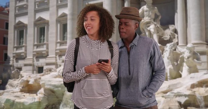 Happy mixed race woman with her boyfriend using smartphone by the Trevi fountain, Smiling millennial couple standing outdoors in Rome, 4k