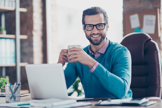 Stunning, smiling,  manly IT-manager in glasses, pullover, shirt, sitting on leather chair at desktop in workplace, looking at camera, holding mug with tea