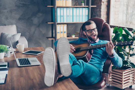 Cheerful, creative man sitting at desktop on chair, putting legs on table, having guitar in hands, playing song and singing in work place