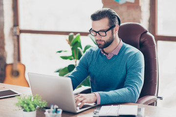 Stunning man in shirt and pullover sitting on leather chair at desktop in work station, with concentrated expression looking at screen of laptop, searching information, works on project, presentation