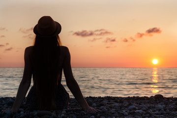 Girl in the hat sitting on the seashore. Sunset time. Silhouette.  Back view