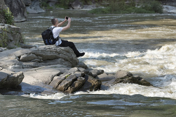 Young man on the bank of a stormy river takes a photo on a smartphone. Sunny summer day